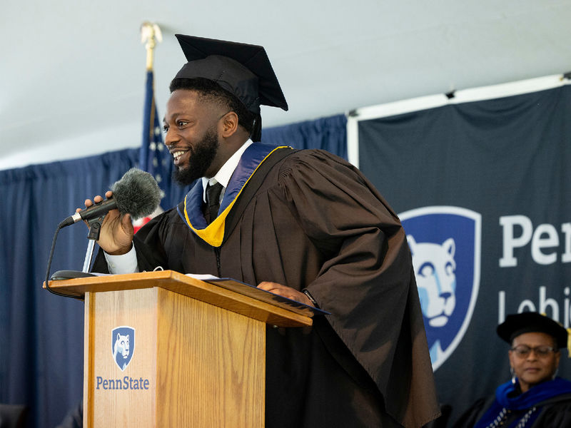 Abu Fofanah wearing commencement regalia standing at a podium speaking.