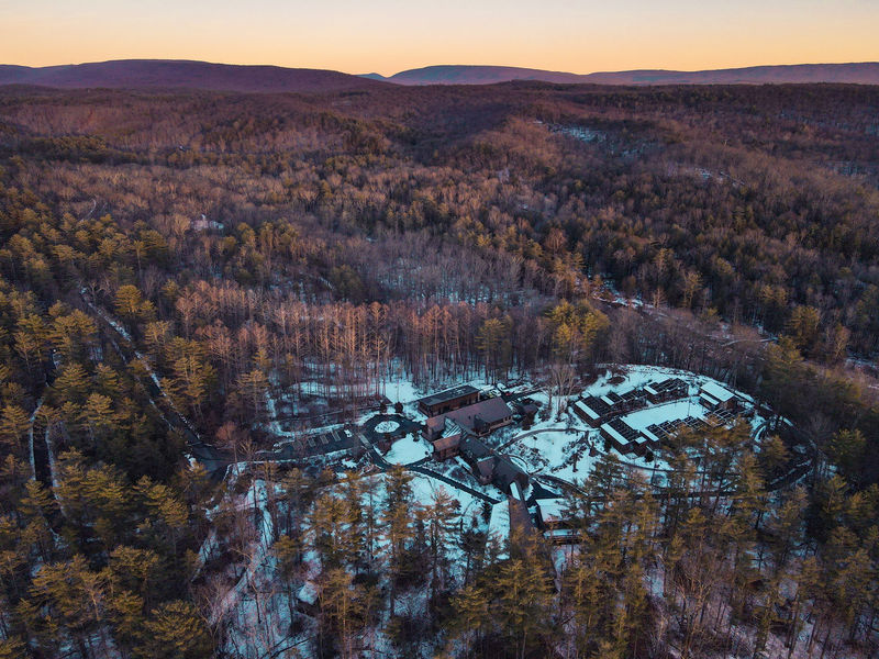 An aerial photograph of a mountainside lined with trees with brown leaves, with a small patch of snowy ground in the center