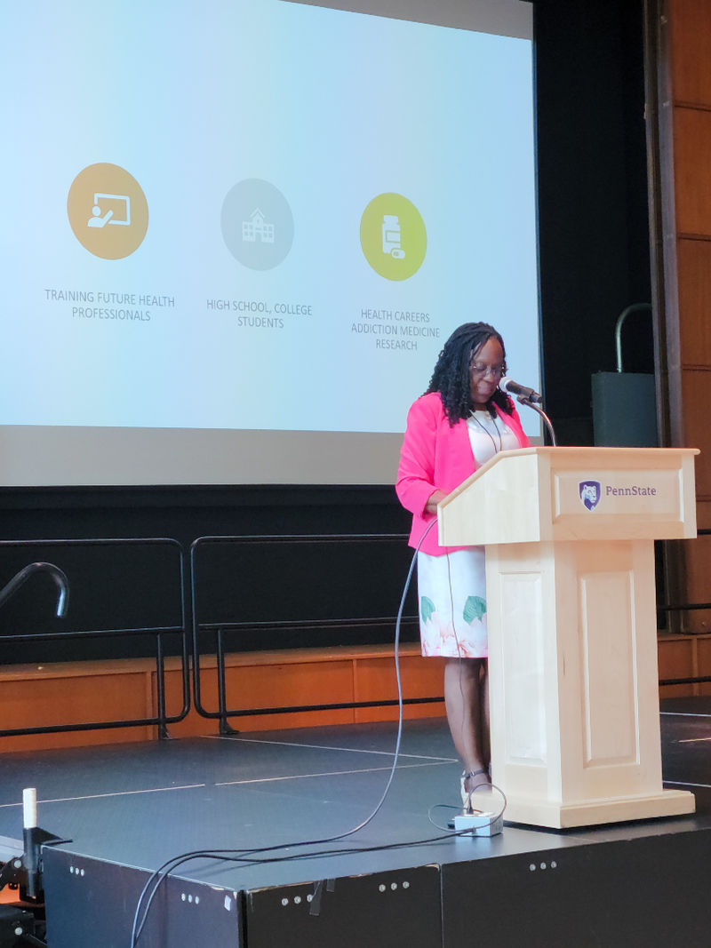 Olapeju Simoyan, a black woman wearing a bright pink blazer, stands at a podium.