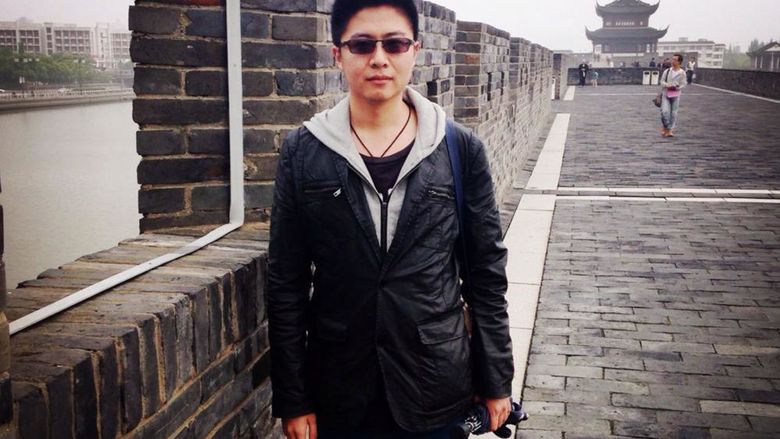 A person in sunglasses and a light jacket poses in front of a brick wall on a gray day. Buildings with traditional Chinese and with modern architecture can be seen in the background. 