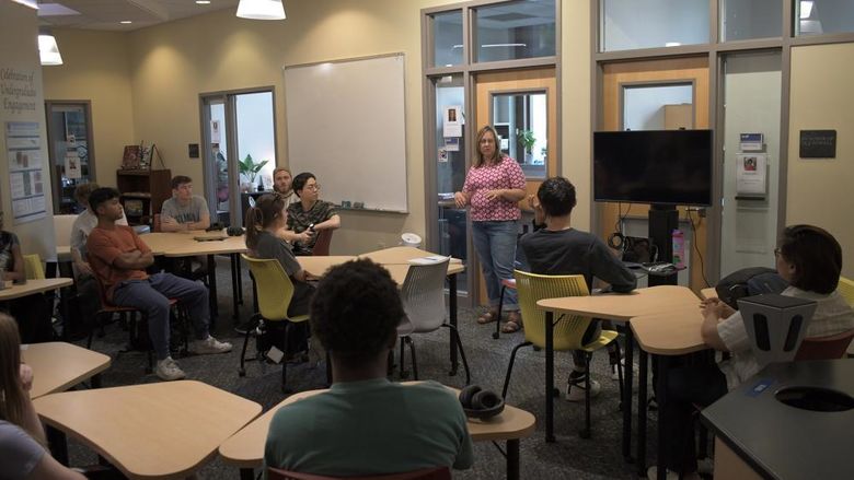 Person stands at the front of a room, talking to other people sitting at tables. 