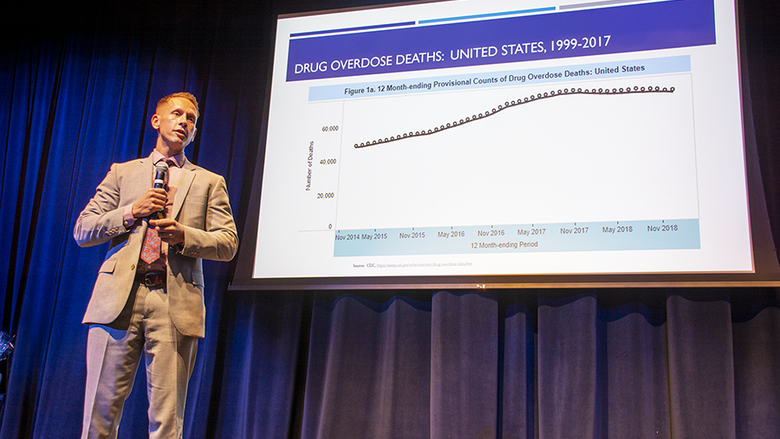 Dr. Glenn Sterner stands on stage as he delivers a speech. There is a slide behind him reviewing drug overdose deaths in the U.S. from 1999-2017 