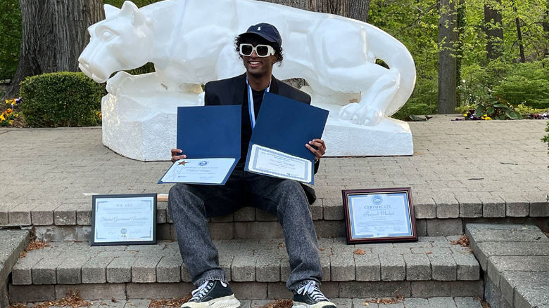 Student in sunglasses poses with awards in front of white Nittany Lion shrine 