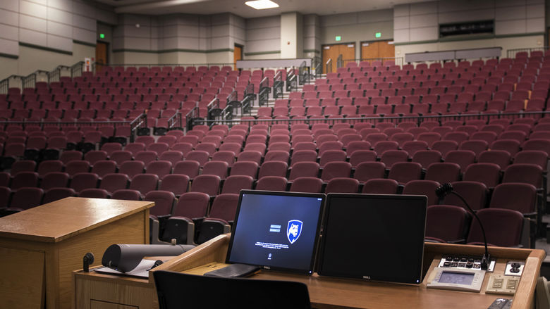 A computer screen is lit up at a podium overlooking an empty theater classroom.