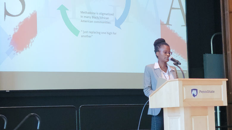 Imani Brown, a black woman in a grey blazer and black pants, stands at the podium.