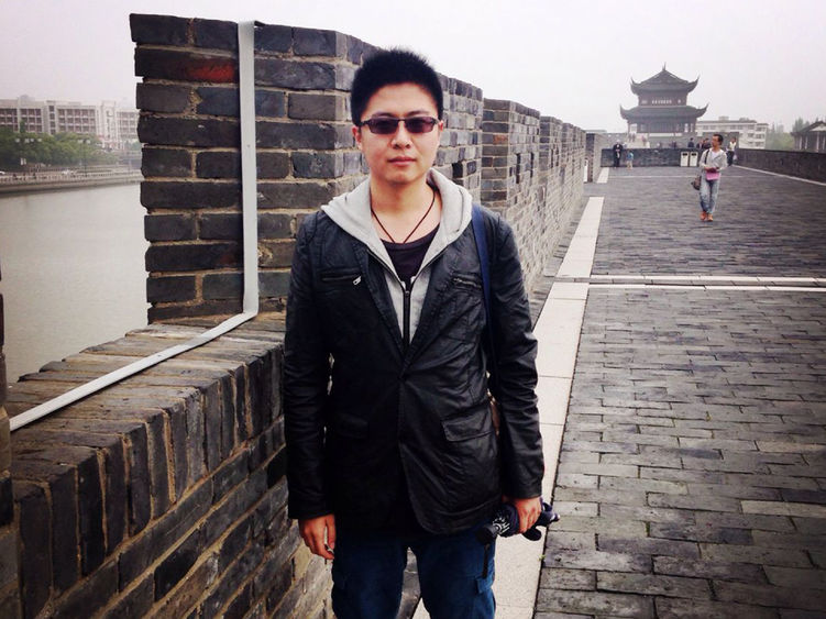 A person in sunglasses and a light jacket poses in front of a brick wall on a gray day. Buildings with traditional Chinese and with modern architecture can be seen in the background.